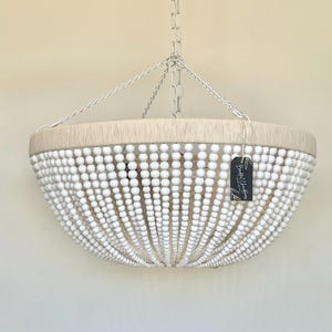 May include: A bowl-shaped pendant light fixture featuring a frame wrapped in light beige fiber, adorned with numerous vertical strands of small white beads. The fixture is suspended by a silver-toned metal chain and includes a small black tag.
