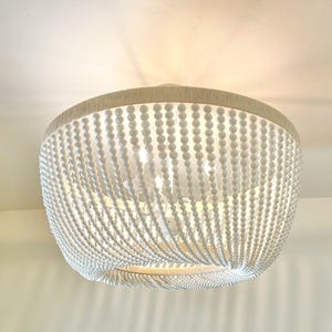 May include: A white beaded flush mount ceiling light fixture. The light has a round shape with multiple strands of white beads hanging from the top. The top of the fixture is wrapped in a beige fabric.