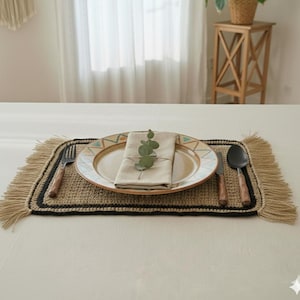 Peut inclure: Une mise en place avec un set de table en jute tressé, bordure noire et franges. Une assiette en céramique avec un motif géométrique contient une serviette en lin pliée et une branche d'eucalyptus. Couverts avec des manches en bois.