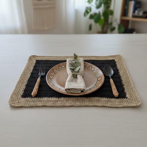May include: A placemat with a black center and a tan crocheted border. A tan plate with a geometric design, a folded napkin, and silverware with wooden handles are arranged on the placemat. A sprig of greenery is on the napkin.