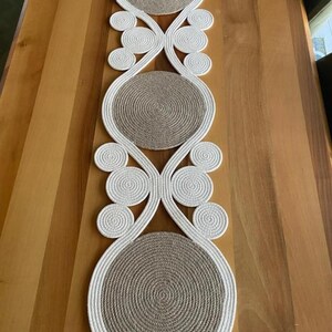 May include: A white and brown woven table runner with a decorative design. The runner has a circular pattern with a white rope border and a brown center.