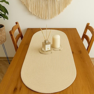 May include: A beige oval table runner with a textured, ribbed design, placed on a wooden table. A candle, a diffuser, and coasters are on the runner. Two wooden chairs are on either side of the table. A macrame wall hanging is in the background.