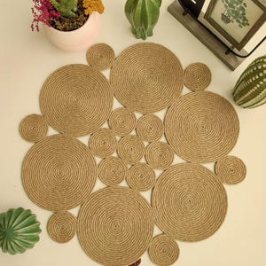 Puede incluir: Un mantel individual redondo de yute tejido beige con un diseño único de círculos superpuestos. El mantel individual está hecho de fibras naturales y tiene un aspecto rústico y terroso.