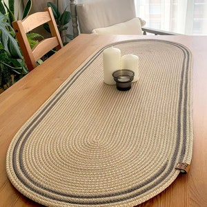 Op de afbeelding: Een beige ovale tafelrunner met een grijze gevlochten rand. De runner is gemaakt van geweven materiaal en heeft een subtiele textuur. Twee witte kaarsen en een kaarsenhouder zijn op de runner geplaatst.