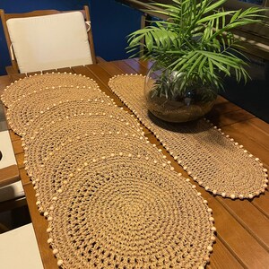 May include: Two oval-shaped crocheted placemats with white beads around the edges. The placemats are made of natural fiber and are on a wooden table.