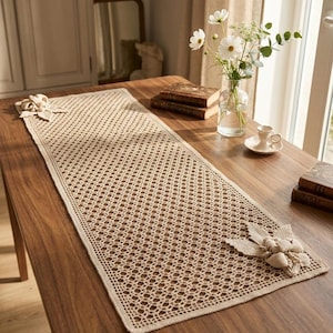 May include: A rectangular beige crochet table runner featuring an intricate openwork diamond pattern throughout the center. The runner is finished with a solid border and large, three-dimensional crocheted floral embellishments at two corners.