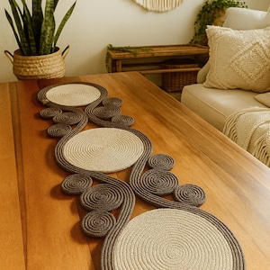 Può includere: Runner da tavolo decorativo in corda beige e marrone. Il runner presenta tre ampie aree circolari per sottopiatti collegate da motivi a spirale in corda. È posizionato su un tavolo di legno.
