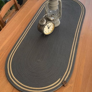 May include: Oval gray braided table runner with a tan border. A vintage alarm clock and a gray lantern sit on the runner, which is placed on a wooden table. The runner is approximately 36 inches long.