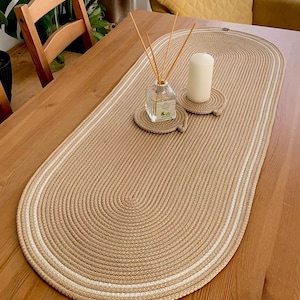 May include: A beige oval table runner with a white border. The runner is made of a woven material and has a textured surface. The runner is on a wooden table with a candle and a diffuser on top.