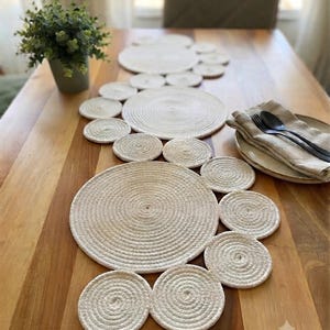 May include: A table runner composed of multiple off-white circular rope mats of varying sizes arranged in a cluster. The mats feature a coiled design with a textured surface, creating a continuous decorative piece for a dining table surface.