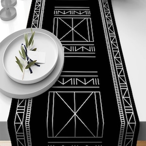 May include: Black and white geometric table runner with a repeating pattern of squares and triangles. The runner has a white border and is made of a soft, durable fabric.