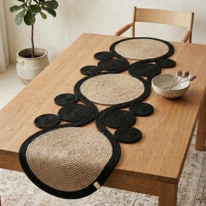 May include: A natural fiber table runner with a unique design. The runner features large, circular placemats in a light tan color with a black border, connected by smaller black circles. The runner is placed on a wooden table.