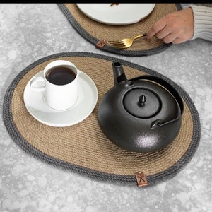 May include: Two oval jute placemats with a grey trim. The placemats are made of natural jute fibre and have a rustic look. One placemat has a black teapot and a white coffee cup with a saucer on it. The other placemat has a gold fork on it.