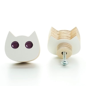 Może przedstawiać: Dwa białe drewniane uchwyty do szuflad w kształcie kota z fioletowymi szklanymi oczami. Jeden uchwyt jest pokazany z przodu, a drugi z boku, ukazując śrubę i elementy montażowe.