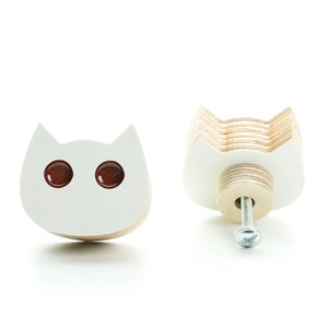 Puede incluir: Tirador de cajón de madera blanco en forma de gato con ojos marrones y un tornillo plateado. El tirador se muestra de frente y de lado.