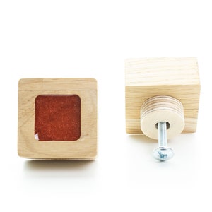 Puede incluir: Dos pomos de armario cuadrados de madera. Un pomo tiene un inserto cuadrado rojo y el otro tiene un tornillo plateado.