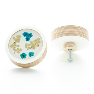 May include: A round wooden knob with a white painted surface and a clear resin insert featuring pressed blue and yellow flowers. The knob has a silver screw for attaching to a drawer or door.