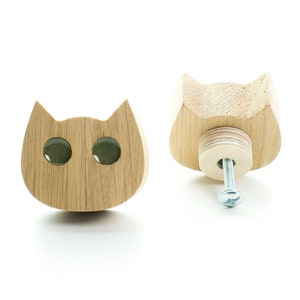 May include: Two wooden cat-shaped drawer pulls with green eyes. The pulls are made of light wood and have a screw on the back.