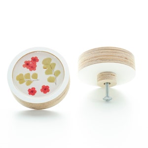May include: A round wooden knob with a white centre featuring pressed red flowers and green leaves. The knob has a silver screw for attaching to a drawer or door.