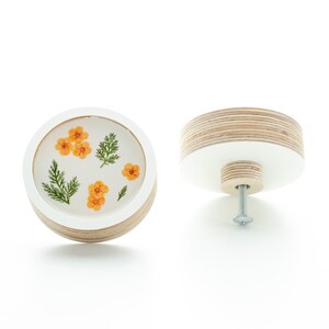 Op de afbeelding: Ronde, witte houten knop met een transparante harsinleg met geperste oranje bloemen en groene bladeren.