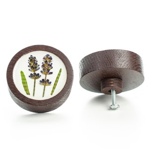 Peut inclure: Un bouton rond en bois avec un fond blanc et un motif de fleur de lavande pressée. Le bouton est brun et a une vis au dos.