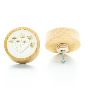 Op de afbeelding: Een ronde houten knop met een wit centrum met een geperst gedroogd bloemenontwerp. De knop wordt vanuit twee hoeken getoond, één met het bloemenontwerp en de andere met de achterkant met een schroef.