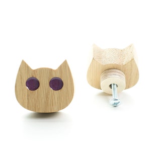 Pode incluir: Dois puxadores de gaveta de madeira em forma de gato com olhos roxos. Os puxadores são feitos de madeira clara e têm um parafuso na parte de trás.