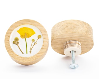 Op de afbeelding: Twee ronde houten knoppen, waarvan er één een geperste gele bloem aan de voorkant heeft. De knoppen hebben een schroef aan de achterkant om ze aan een lade of kast te bevestigen.
