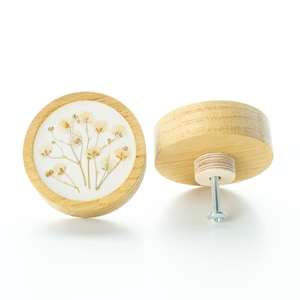 Op de afbeelding: Twee ronde houten knoppen met een witte achtergrond en gedroogde bloemen. Eén knop heeft een schroef aan de onderkant.