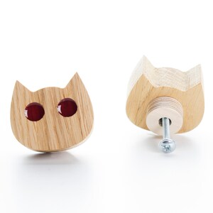 Può includere: Una maniglia per cassetto in legno a forma di gatto con occhi di vetro rosso. La maniglia è mostrata dal davanti e dal dietro.