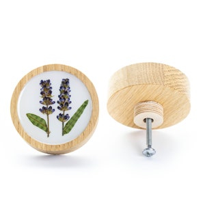 May include: A round wooden knob with a white background and a pressed lavender flower design. The knob has a silver screw and a wooden base.