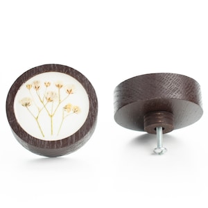 Op de afbeelding: Een ronde, donkerbruine houten knop met een wit middenstuk met een geperst gedroogd bloemenontwerp. De knop heeft een zilveren schroef aan de achterkant.