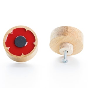May include: Two wooden cabinet knobs. One knob features a red poppy design with a black center. The other knob is a plain wooden cylinder with a screw for installation. The knobs are made of light-colored wood.