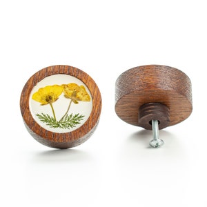 May include: A pair of wooden drawer knobs with a round, flat design. One knob features a pressed, clear resin insert with a dried yellow flower pressed inside. The other knob shows the back side of the knob with a screw.