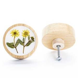Może przedstawiać: Okrągły drewniany gałka z białym tłem i wzorem z prasowanych kwiatów z dwoma żółtymi kwiatami i zielonymi liśćmi. Gałka ma srebrną śrubę z tyłu.