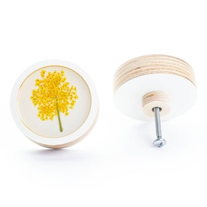 Peut inclure: Bouton rond en bois blanc avec un insert en résine transparente présentant une fleur jaune pressée. Le bouton est doté d'une vis argentée pour la fixation sur une surface.