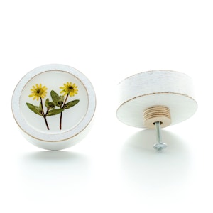 Può includere: Pomo rotondo in legno bianco con un inserto in resina trasparente che presenta due fiori gialli pressati. Il pomo ha una vite bianca e una base in legno.