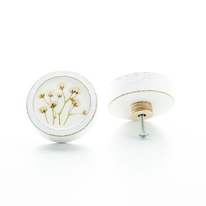 Könnte beinhalten: Runder weißer Schrankknopf mit einem gepressten Blumendesign. Der Knopf hat eine Holzbasis und eine Metallschraube.