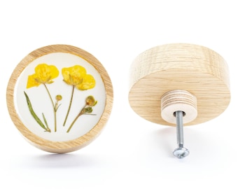 Op de afbeelding: Een ronde houten knop met een transparante harsinleg met geperste gele bloemen. De knop heeft een zilveren schroef om te bevestigen aan een lade of kast.