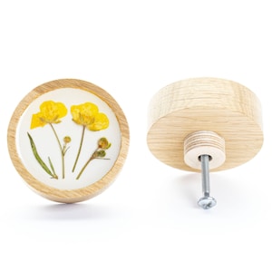 Può includere: Un pomello rotondo in legno con un inserto in resina trasparente che presenta fiori gialli pressati. Il pomello ha una vite argentata per il fissaggio a un cassetto o un armadio.