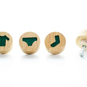 May include: Four wooden knobs with green accents. Each knob features a different clothing item: a t-shirt, underwear, and a sock. The fourth knob is shown with a screw.