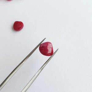 May include: A deep red gemstone with a faceted heart shape, held by tweezers against a white background.