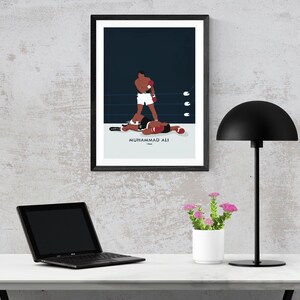 May include: A black framed poster with a minimalist illustration of Muhammad Ali in a boxing match. The poster is hanging on a gray wall above a white desk with a laptop, a lamp, and a small potted plant.