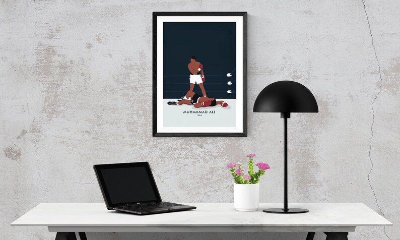 May include: A black framed poster with a minimalist illustration of Muhammad Ali in a boxing match. The poster is hanging on a gray wall above a white desk with a laptop, a lamp, and a small potted plant.