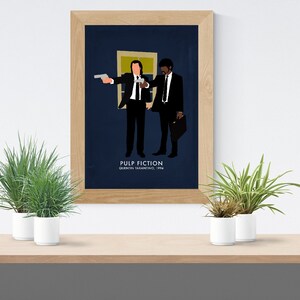 May include: A framed art print featuring a minimalist illustration of two men in black suits, one holding a gun. The background is dark blue. The text "PULP FICTION" and "QUENTIN TARANTINO, 1994" are at the bottom. The frame is light brown.