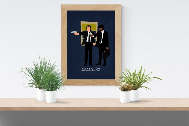 May include: A framed art print featuring a minimalist illustration of two men in black suits, one holding a gun. The background is dark blue. The text "PULP FICTION" and "QUENTIN TARANTINO, 1994" are at the bottom. The frame is light brown.