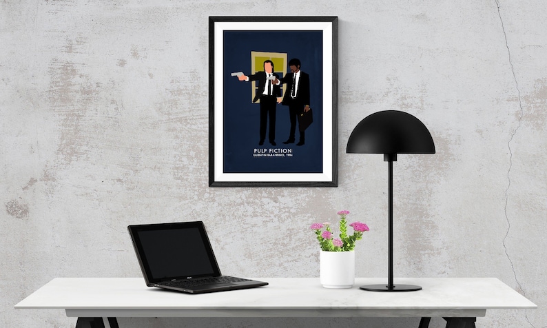 May include: A framed art print featuring a minimalist illustration of two figures from the movie Pulp Fiction. The artwork is set against a dark blue background, with the film's title and director's name at the bottom. The print is displayed on a white desk.