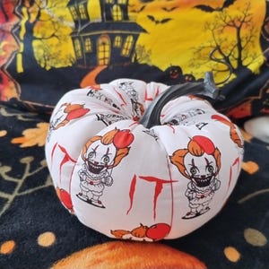 May include: A white fabric pumpkin with a black stem, featuring a repeating pattern of a cartoon clown and the word "IT" in red. The pumpkin is a Halloween decoration.