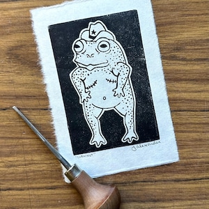 May include: Black and white linocut print of a frog wearing a cowboy hat with a star. The frog has a dotted pattern and is standing with its arms at its sides. The print is on textured paper. The text "CORN NUT" is at the bottom.