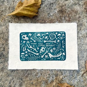 May include: A teal and white rectangular art print featuring various bone and feather illustrations. The text "WHAT WILL YOU LEAVE BEHIND?" is printed in white. The print is on textured white paper, with a stone background and a dried leaf.